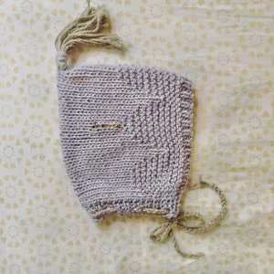 May include: A light purple knitted baby bonnet with a tassel on top and a tie closure. The bonnet has a textured pattern and a small opening near the face. The background is a light yellow fabric with a heart pattern.