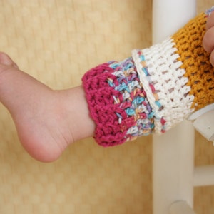 May include: Close-up of a baby's foot and leg wearing colorful, crocheted leg warmers. The leg warmers feature a mix of colors including pink, white, yellow, and blue. The baby's hand is visible.