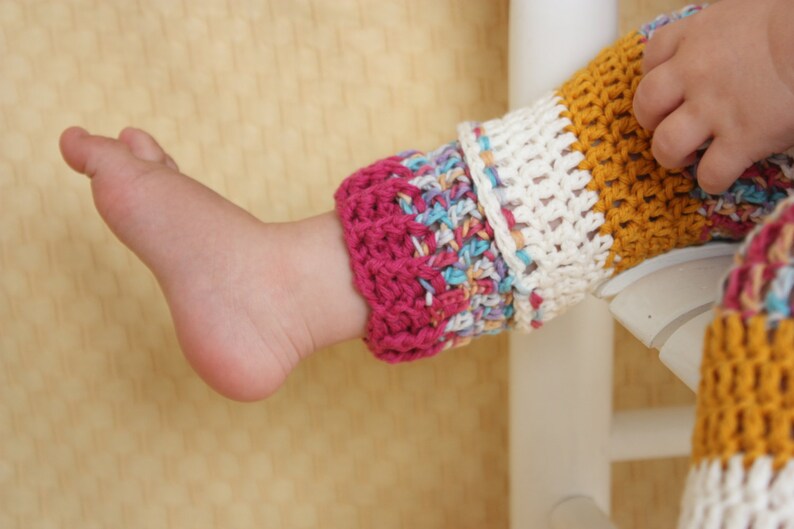 May include: Close-up of a baby's foot and leg wearing colorful, crocheted leg warmers. The leg warmers feature a mix of colors including pink, white, yellow, and blue. The baby's hand is visible.