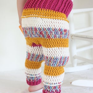 May include: Crocheted baby pants with a ribbed magenta waistband, a mustard yellow band, and striped legs in white, mustard yellow, and multicolored yarn. The pants are handmade and suitable for a baby.