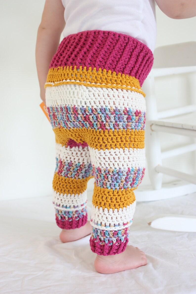 May include: Crocheted baby pants with a ribbed magenta waistband, a mustard yellow band, and striped legs in white, mustard yellow, and multicolored yarn. The pants are handmade and suitable for a baby.