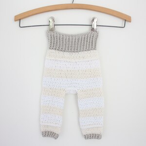 May include: Crocheted baby pants in a striped pattern of white, cream, and gray. The pants are suspended from a wooden hanger with a metal clip. The waistband and cuffs are solid gray.