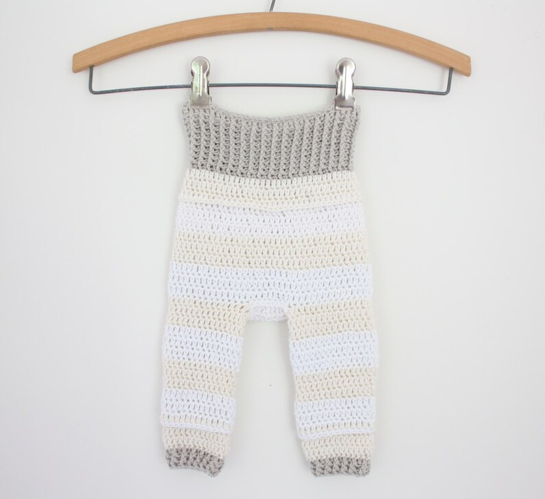 May include: Crocheted baby pants in a striped pattern of white, cream, and gray. The pants are suspended from a wooden hanger with a metal clip. The waistband and cuffs are solid gray.