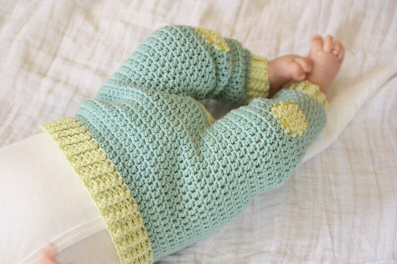 May include: A pair of baby pants crocheted in a light green color with yellow accents. The pants have a textured stitch pattern and are folded at the ankles.