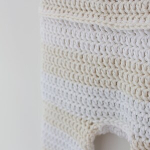 May include: Close-up of a crocheted garment in cream and white. The handmade item features a textured stitch pattern with horizontal stripes of varying shades of cream and white. The garment appears to be a baby item.