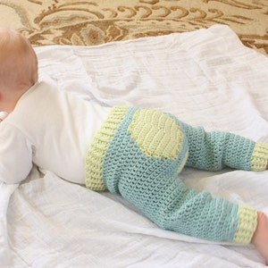 May include: A baby wearing a light blue and yellow crocheted pants with a yellow circle on the back. The baby is lying on a white blanket.