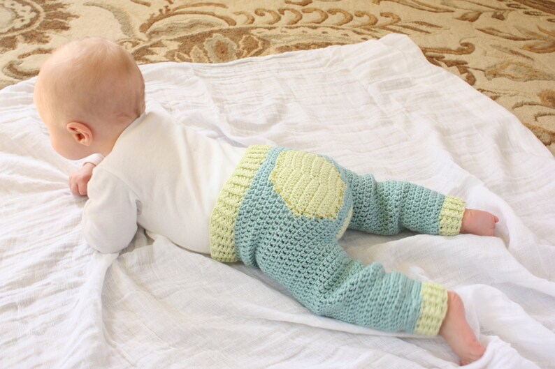 May include: A baby wearing a light blue and yellow crocheted pants with a yellow circle on the back. The baby is lying on a white blanket.