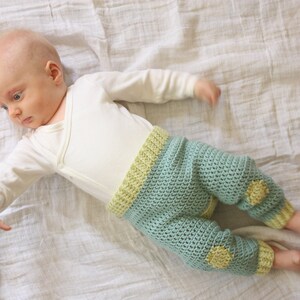 May include: A baby wearing a white long-sleeved onesie and crocheted pants with yellow accents. The pants have a light blue color and are crocheted in a textured pattern.