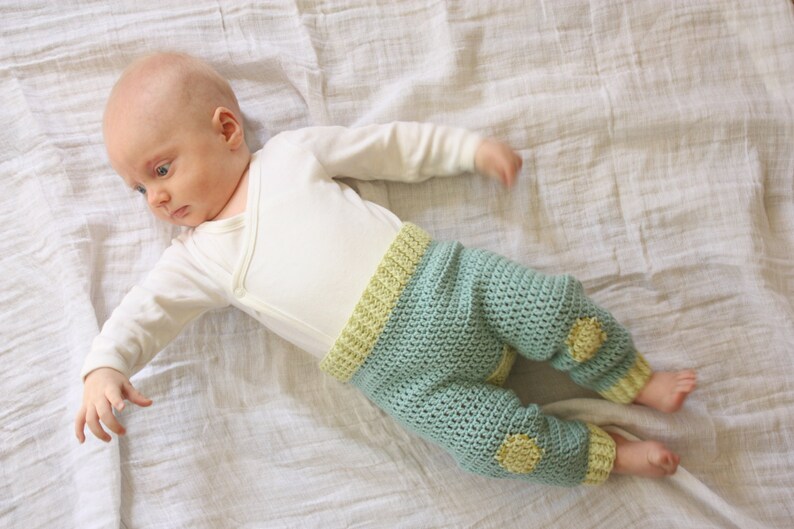 May include: A baby wearing a white long-sleeved onesie and crocheted pants with yellow accents. The pants have a light blue color and are crocheted in a textured pattern.
