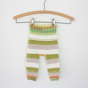 May include: Striped crochet baby pants in green, white, and orange. The pants are hanging on a wooden hanger.