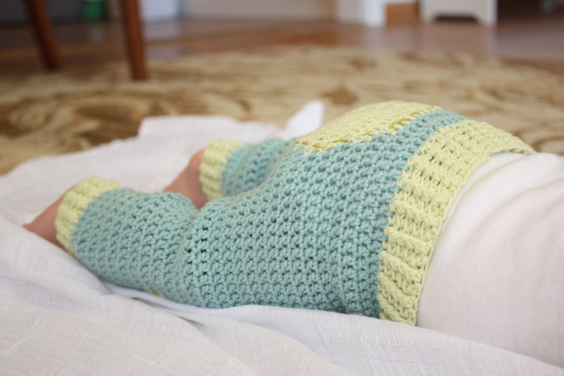 May include: A baby wearing a crocheted outfit with a blue and yellow color scheme. The outfit is made of soft yarn and has a loose fit.