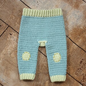 May include: A pair of crocheted baby pants in light blue with yellow accents. The pants have a yellow band at the waist and yellow circles on the knees.