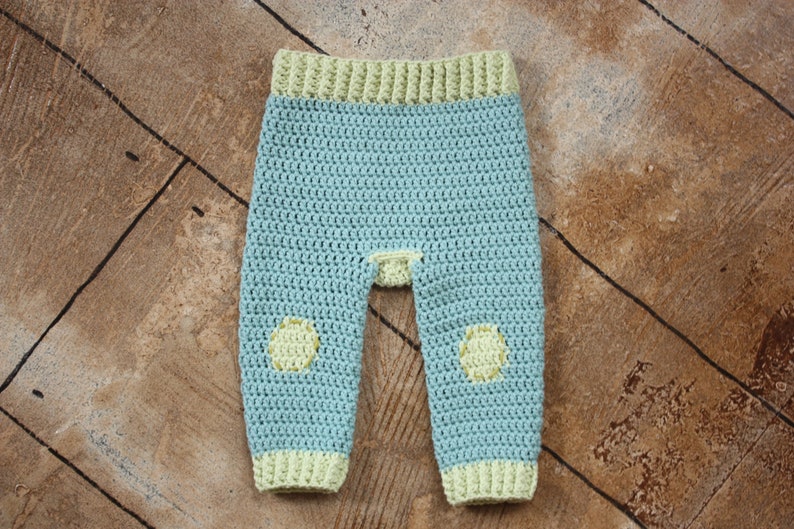 May include: A pair of crocheted baby pants in light blue with yellow accents. The pants have a yellow band at the waist and yellow circles on the knees.