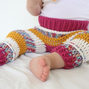 May include: Hand-crocheted baby pants in a colorful design. The pants feature a fuchsia waistband and cuffs, with sections of yellow, white, and multicolored yarn. The open weave design is visible. A comfortable and stylish choice for infants.