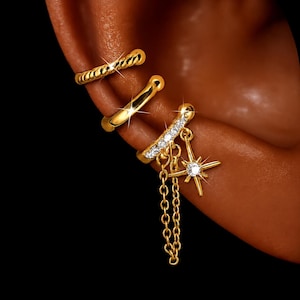 Puede incluir: Un conjunto de tres pendientes tipo ear cuff dorados. El superior tiene una textura trenzada, el central es una banda lisa y el inferior presenta una hilera de piedras claras, un dije en forma de estrella con una piedra central y una cadena fina.