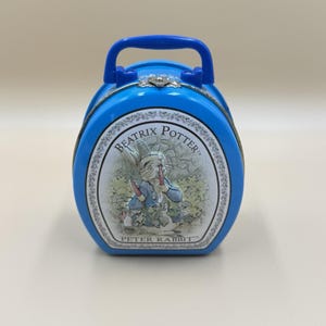 May include: A bright blue oval-shaped carrying case with a top handle and a metal latch. The front features an oval illustration of Peter Rabbit from Beatrix Potter, with the text BEATRIX POTTER and PETER RABBIT printed in black on a white background.