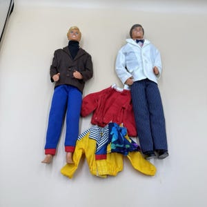 May include: Two male dolls with molded hair, one wearing a brown blazer over a black turtleneck with blue pants, the other in a white blazer with a bowtie and pinstriped pants. Included are a red long-sleeve shirt and a yellow jacket with blue accents.