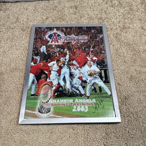 May include: A framed photograph of the 2002 World Champions Anaheim Angels baseball team celebrating on the field. The image features multiple player autographs, the team logo, and the text Anaheim Angels Spring Training 2003.