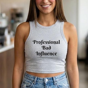 Op de afbeelding: Een lichtgrijze mouwloze crop top met een geribbelde ronde hals. De voorkant van het kledingstuk is voorzien van de zwarte tekst "Professional Bad Influence" gedrukt in een vet, schreeflettertype in het midden van de borst.
