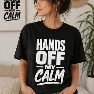 Könnte beinhalten: Ein schwarzes kurzärmeliges T-Shirt mit dem weißen Schriftzug "HANDS OFF MY CALM", der in einer fetten, stilisierten Schrift auf der Vorderseite gedruckt ist. Das Design enthält eine horizontale Linie über dem Wort "CALM" und einen Pinselstrich.
