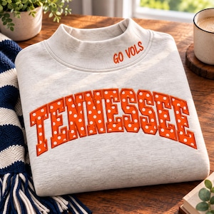 Puede incluir: Una sudadera gris jaspeado claro con cuello alto. El texto GO VOLS está bordado en naranja en el cuello. La palabra TENNESSEE está aplicada en el pecho en letras de bloque naranjas con un patrón de lunares blancos.