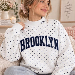Op de afbeelding: Een wit sweatshirt met lange mouwen, een donkerblauw stippenpatroon en het woord BROOKLYN in grote donkerblauwe blokletters op de borst. De opstaande kraag is voorzien van de tekst 95er, geborduurd in een klein donkerblauw cursief lettertype.