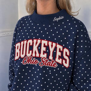 May include: A navy blue sweatshirt with a white polka dot pattern, featuring the embroidered text BUCKEYES Ohio State in red and white on the chest. The name Sophia is embroidered in white cursive script on the ribbed mock neck collar.