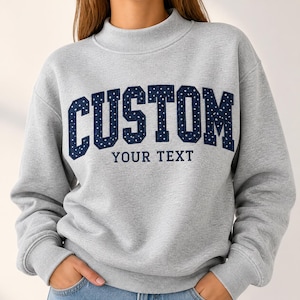 Puede incluir: Una sudadera gris claro de manga larga con cuello alto. La parte delantera presenta la palabra CUSTOM en letras grandes de bloque azul marino oscuro con un patrón de puntos blancos, y las palabras YOUR TEXT en letras azul marino más pequeñas.