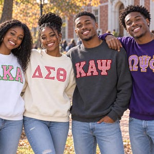 May include: Four crewneck sweatshirts featuring Greek letters in a gingham pattern. The first is white with green letters, the second is cream with red letters, the third is charcoal gray with red letters, and the fourth is purple with purple letters.