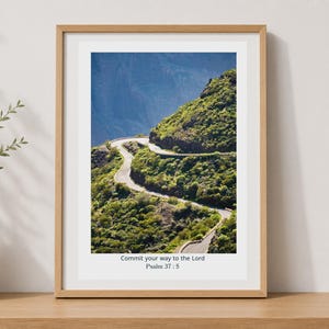 May include: A framed vertical wall art print featuring a winding mountain road surrounded by green vegetation and rocky cliffs. The bottom white border displays the black text: Commit your way to the Lord, Psalm 37:5. The frame is light wood.
