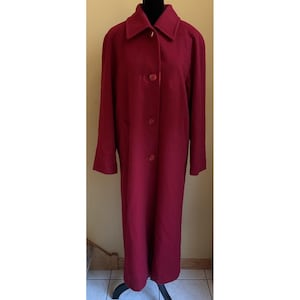 May include: A long, deep red coat featuring a structured collar, long sleeves, and a front button closure with four visible matching buttons. The coat has a straight, full-length silhouette and is displayed on a black mannequin torso.