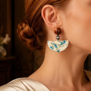 May include: A fan-shaped earring featuring a cream-colored base with a blue floral pattern. The earring is suspended from a smooth, round, dark brown wooden bead, connected by a small, silver-toned cylindrical spacer adorned with clear crystals.