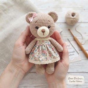 Può includere: Un orsacchiotto all'uncinetto realizzato in filato marrone chiaro con muso color crema e occhi neri. L'orso indossa un vestito con stampa floreale rosa e blu, un colletto in pizzo color crema e un piccolo fiocco rosa sull'orecchio destro.