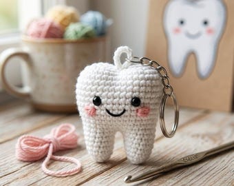 May include: A white crocheted tooth keychain featuring black bead eyes, a stitched black smile, and pink embroidered cheeks. The plush tooth has a metal keyring attached to the top. A silver crochet hook and pink yarn are placed nearby.