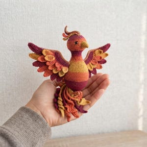 Könnte beinhalten: Eine gehäkelte Phönix-Figur mit einem Farbverlauf aus tiefem Violett, leuchtendem Orange und Goldgelb. Der Vogel hat ausgebreitete Flügel mit geschichteten Federdetails, einen kleinen Kamm auf dem Kopf und einen langen, fließenden Schwanz.