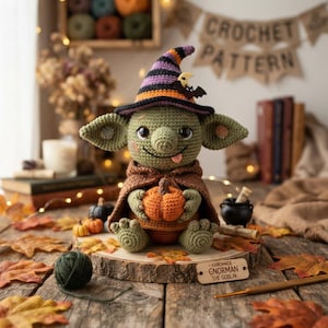 May include: A crocheted green goblin doll with large ears, wearing a purple, orange, and black striped witch hat with a moon and bat accent. The goblin wears a brown cape, holds a small orange pumpkin, and sits on a wooden base with a tag reading HANDMADE GNORMAN THE GOBLIN.