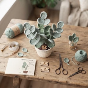 May include: A crocheted eucalyptus plant in a white pot, accompanied by two balls of light green yarn, two pairs of metal scissors, a measuring tape, two wooden tags labelled Eucalyptus, and a printed instruction card titled Crochet Eucalyptus Pattern.