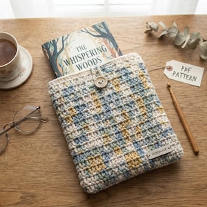 May include: A rectangular crochet sleeve featuring a variegated pattern of cream, light blue, and mustard yellow yarn. The sleeve has a single wooden button closure at the top center and a small rectangular tag labeled "HANDMADE" at the bottom right corner.