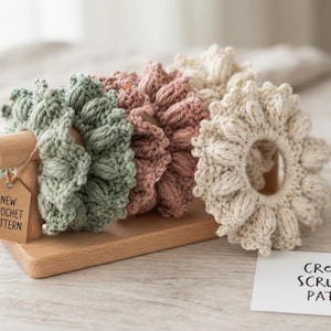 May include: Three crocheted hair scrunchies in sage green, dusty rose, and cream, featuring a textured bobble stitch design. A wooden stand holds the scrunchies with a tag reading NEW CROCHET PATTERN. A card reads CROCHET SCRUNCHIE PATTERN.