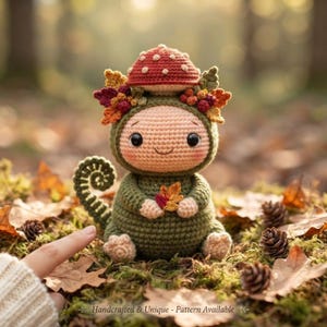 May include: A crocheted creature with a light peach face, large black eyes, and a green body. It wears a red mushroom cap hat adorned with colorful autumn leaves and holds a small orange leaf. It has a curled tail and text at the bottom.