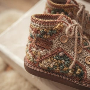 Puede incluir: Un par de botines de ganchillo de caña alta con un patrón geométrico multicolor en tonos marrón, verde y crema. Los botines tienen una etiqueta de cuero marrón con la inscripción HANDMADE COZY STEPS, botones de madera y cordones de hilo.