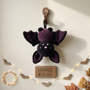 May include: A plush crochet bat with a black body and dark purple wings, hanging from a wooden wall hook. The bat features two black eyes and a small pink nose. Below the bat is a rectangular wooden sign with the text HANDMADE WITH LOVE.