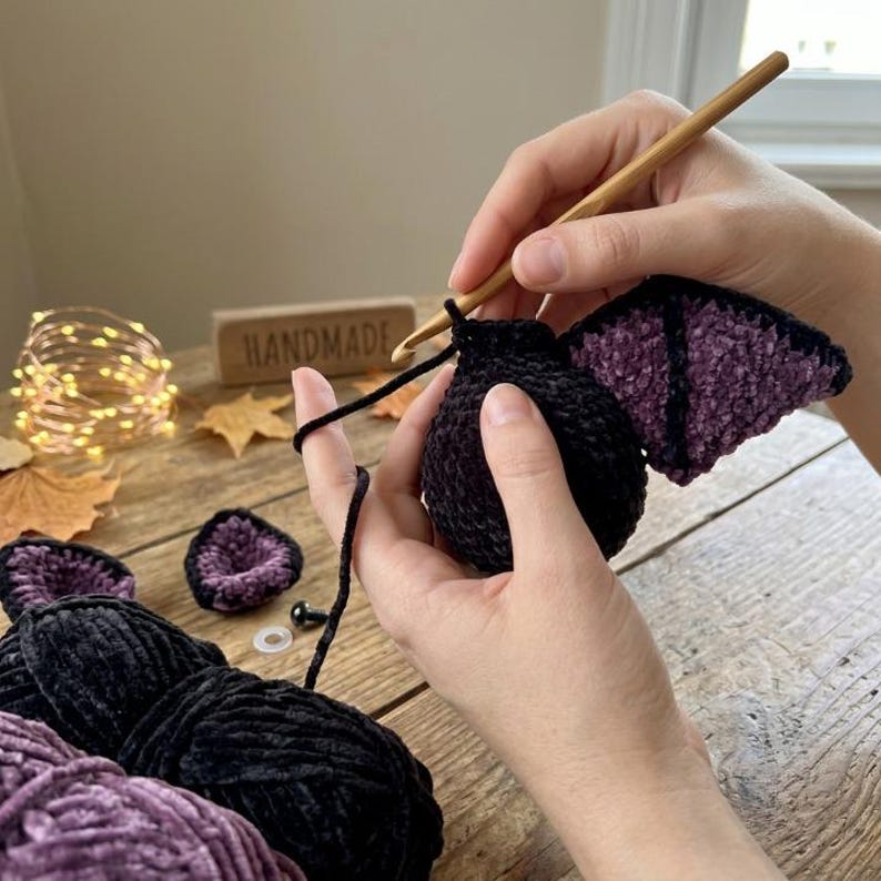May include: A wooden crochet hook is used to work with black and purple velvet yarn to create a plush bat toy. The project includes a round black body, purple-accented wings, and separate small purple and black ear pieces on a wooden surface.