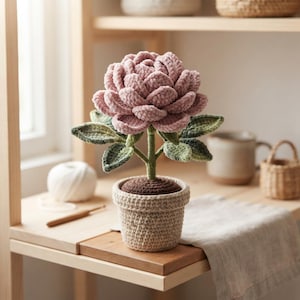 May include: A crocheted flower with layered dusty pink petals and green leaves, potted in a beige textured crochet container with a dark brown soil-like top. The plant features a central green stem and is crafted from soft yarn materials.