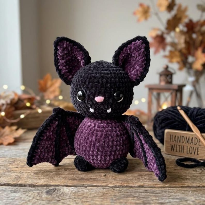 May include: A plush bat toy made of dark purple and black velvet yarn. The bat features large pointed ears, two black plastic eyes, a small pink nose, and two white fangs. Its wings are spread out, and its body is round and textured.