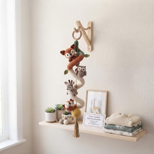May include: A hanging crochet mobile featuring a spiral structure with stuffed woodland animals including a fox, owl, and deer. The mobile is suspended from a wooden wall bracket and ends with a yellow sphere and a multi-colored yarn tassel.