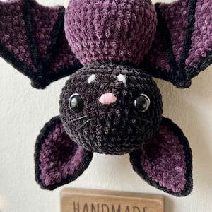 May include: A plush bat toy crafted from dark purple and black yarn. The creature features a round head with large black plastic eyes, a small pink nose, and embroidered whiskers. Its wings are wide and textured with black trim edges.