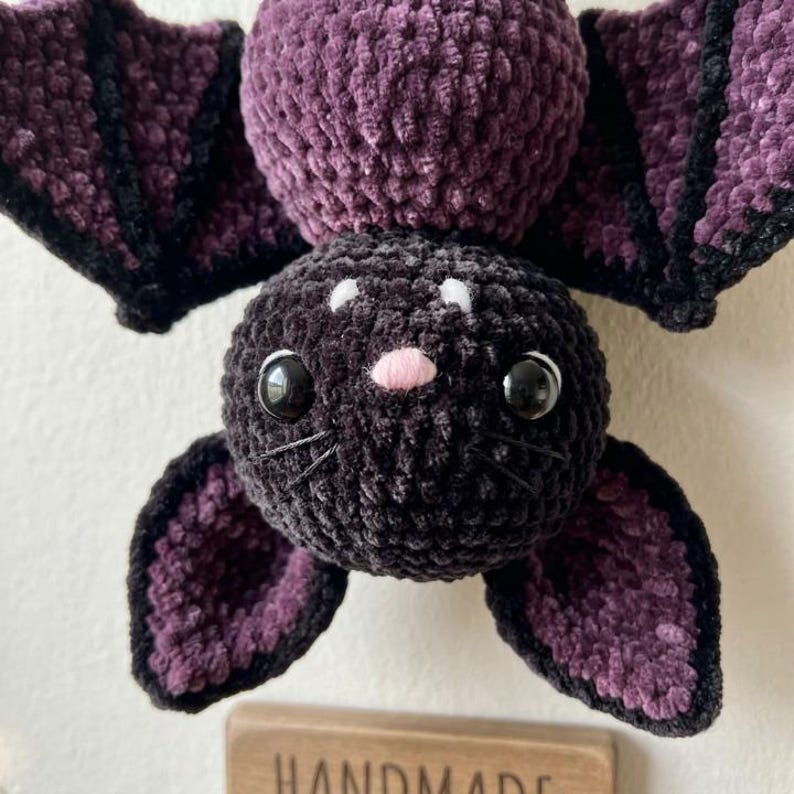 May include: A plush bat toy crafted from dark purple and black yarn. The creature features a round head with large black plastic eyes, a small pink nose, and embroidered whiskers. Its wings are wide and textured with black trim edges.