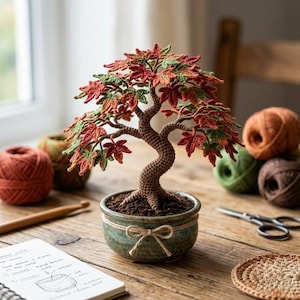 Op de afbeelding: Een gehaakte bonsaiboom met een getextureerde bruine stam en levendige herfstkleurige bladeren in tinten rood, oranje en groen. De boom staat in een kleine, ronde, gespikkelde groene keramische pot, versierd met een eenvoudig gestrikt touwtje.