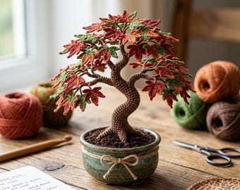 May include: A crocheted bonsai tree with a textured brown trunk and vibrant autumn-colored leaves in shades of red, orange, and green. The tree is planted in a small, round, speckled green ceramic pot decorated with a simple tied twine bow.